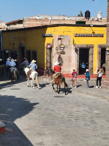 Cowboys riding past pedestrians and storefronts on a sunny San Miguel street